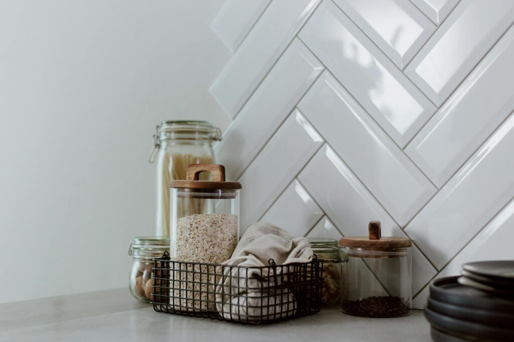 Kitchen counter organized with glass storage jars with wooden lids, a black wire basket, and stacked dark bowls against white herringbone tile backsplash